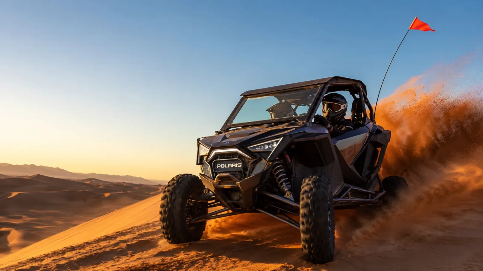 A Polaris RZR sport side-by-side carving across an orange desert dune at golden hour with a rooster tail of dust and a whip flag