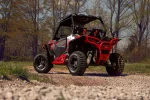 Polaris RZR with red tube rock sliders sitting on gravel in front of bare woods