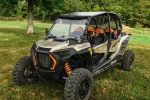 Black aluminum roof installed on a tan four-seat Polaris RZR parked in a grassy clearing at the edge of the trees